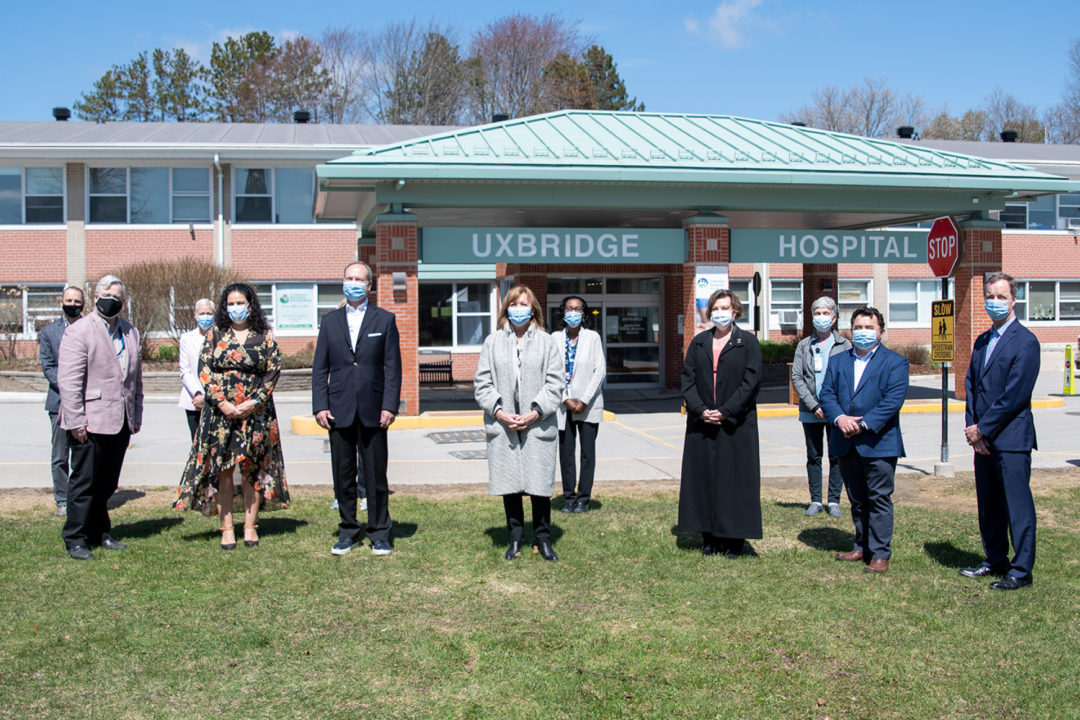 members of the board stand in front of uxbridge hospital's entrance in rows with their hands clasped and wearing masks