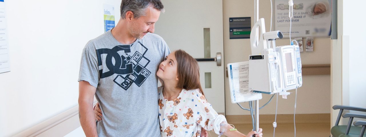 a young girl wearing teddy bear pajamas hugs her father, who wears a grey t-shirt, with one arm while holding onto her IV stand. Both are looking at eachother and smiling