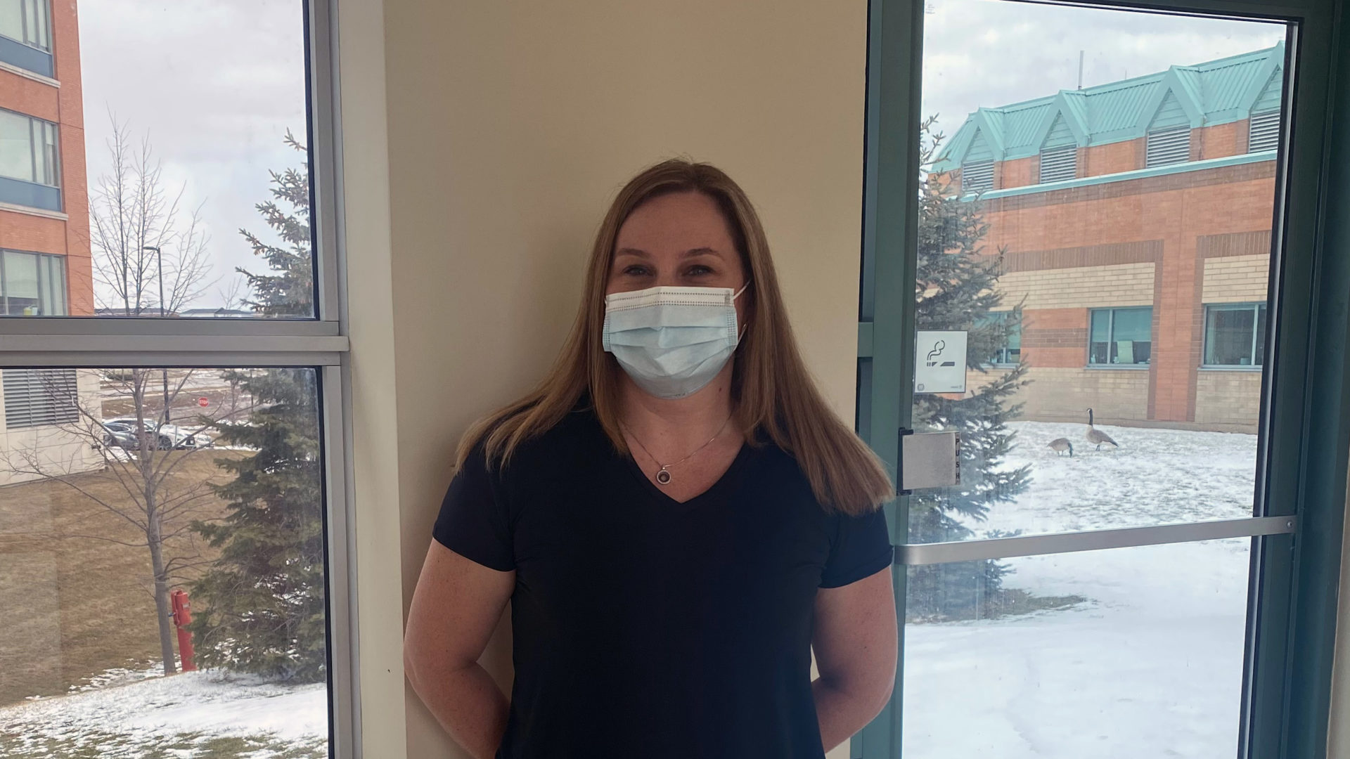 Cindy Stortz, standing against a wall in the hospital, wearing a black shirt, medical mask, and medium length blond hair