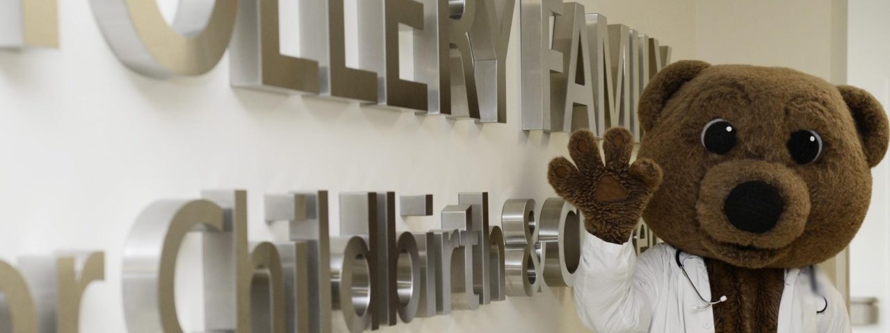 Dr. Bear stands in front of the Stollery family sign and waves for the camera