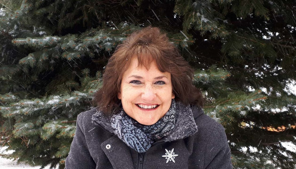 Esther Herbert, wearing a grey coat and short red hair, standing in front of an evergreen tree in the snow