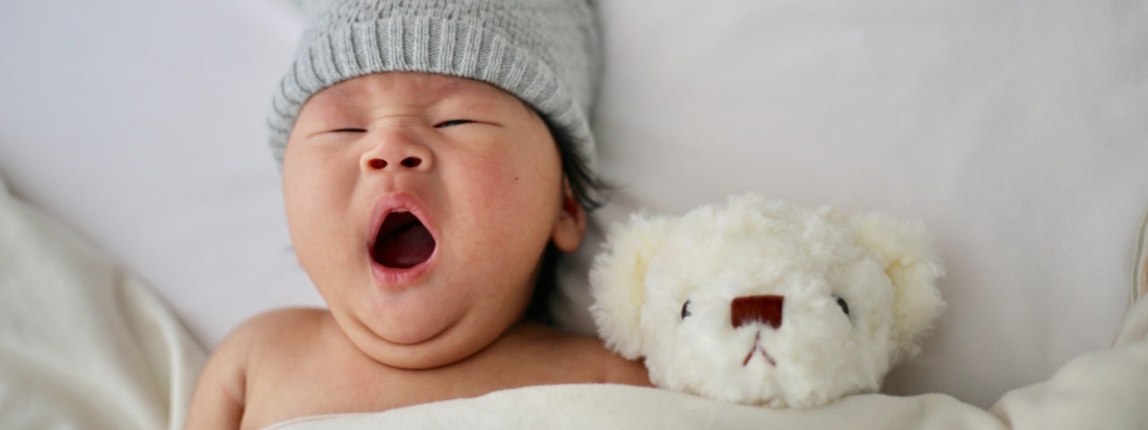 a newborn baby seen from the shoulders up while laying in bed. The baby is yawning and wearing a grey had. A white teddy bear is next to the baby