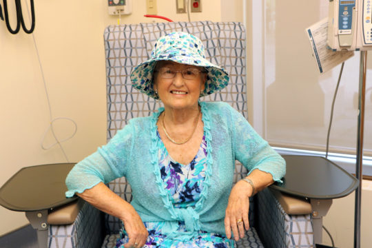 Gerry Clarkson, sitting in a hospital chair, wearing a patterned blue dress and hat, smiling