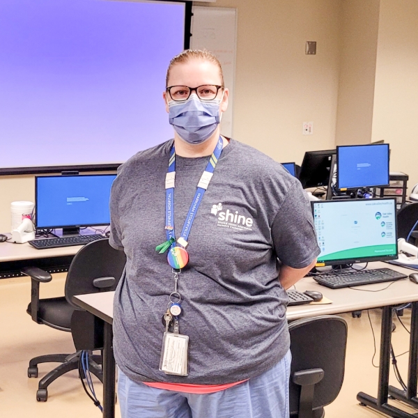 Kelsey Missons standing in front of a row of computers, wearing a gray t-shirt and medical mask