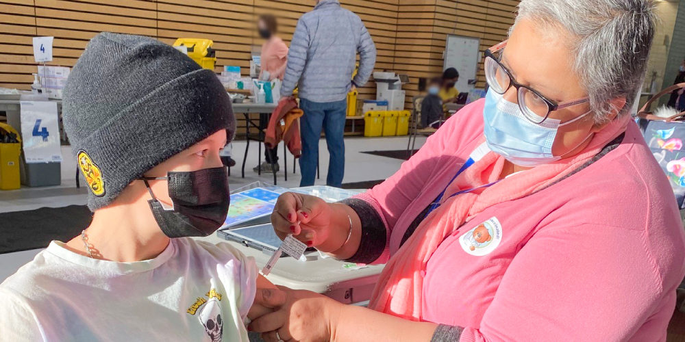 a nurse in a pink cardigan and medical mask gives an injection to a young boy wearing a black beanie and medical mask
