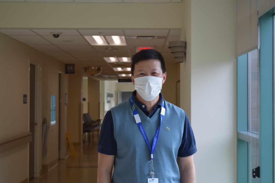 Michael Hunynh wearing a sweater vest, medical mask, and short black hair, poses for a photo in the hospital hallway a