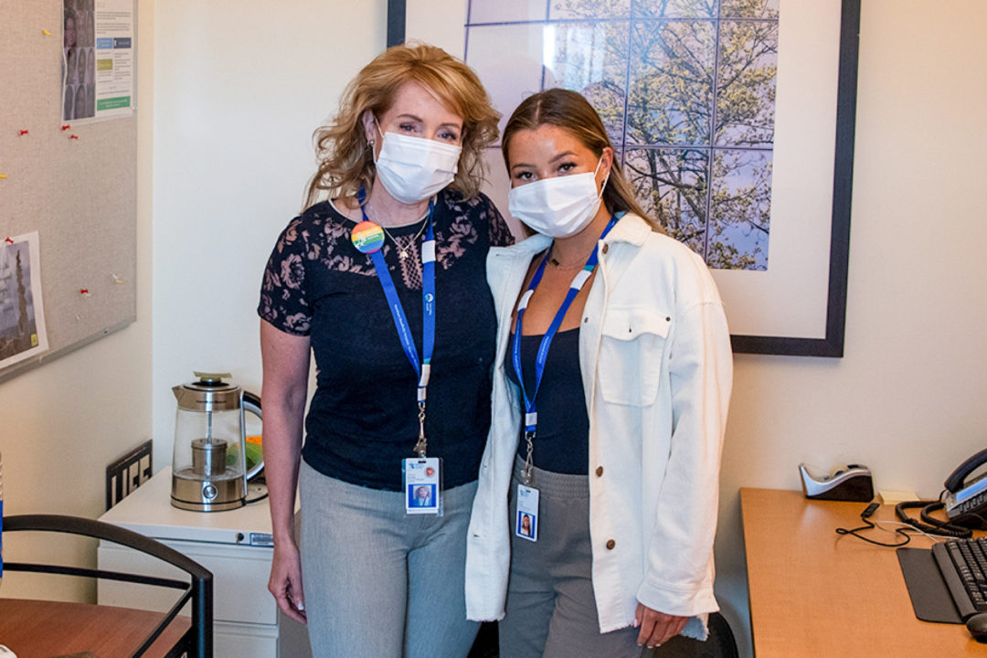 two women in business clothes and wearing medical masks stand together for a photo