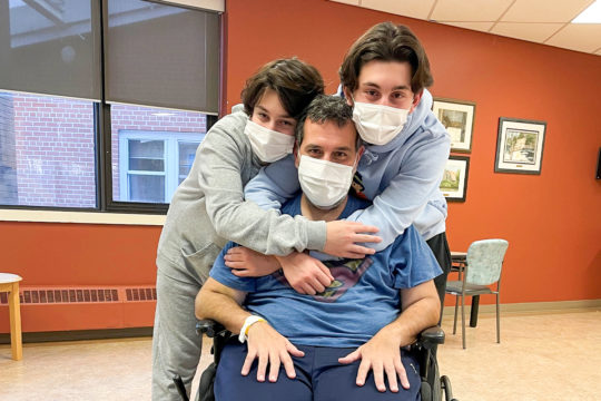 two young boys stand behind their father in a wheel chair and wrap their arms around him