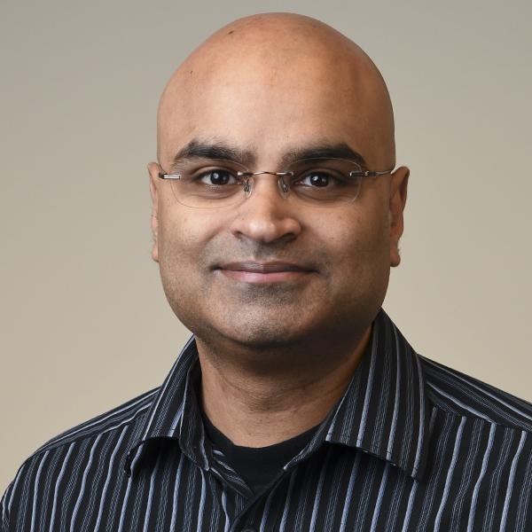 Dr. Sachin Ramkissoon, seen from the chest up, wearing a black and white striped collared shirt, glasses, and smiling