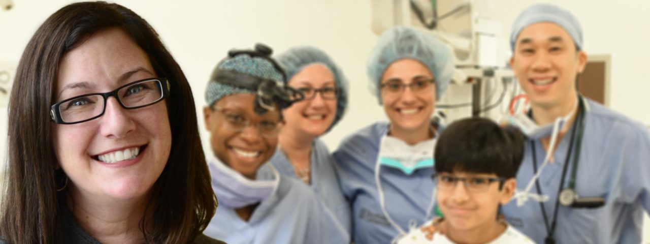 A woman, seen from the neck up and wearing glasses, smiles for the camera while 4 doctors stand around a young patient and smile