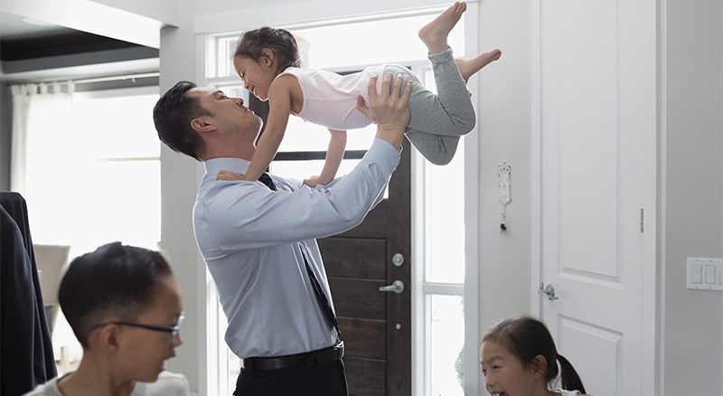 a man in a suit lifts his daughter into the air while his other daughter and son run by him