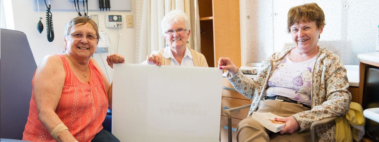 three senior female patients sit together in a hospital room and smile for a photo