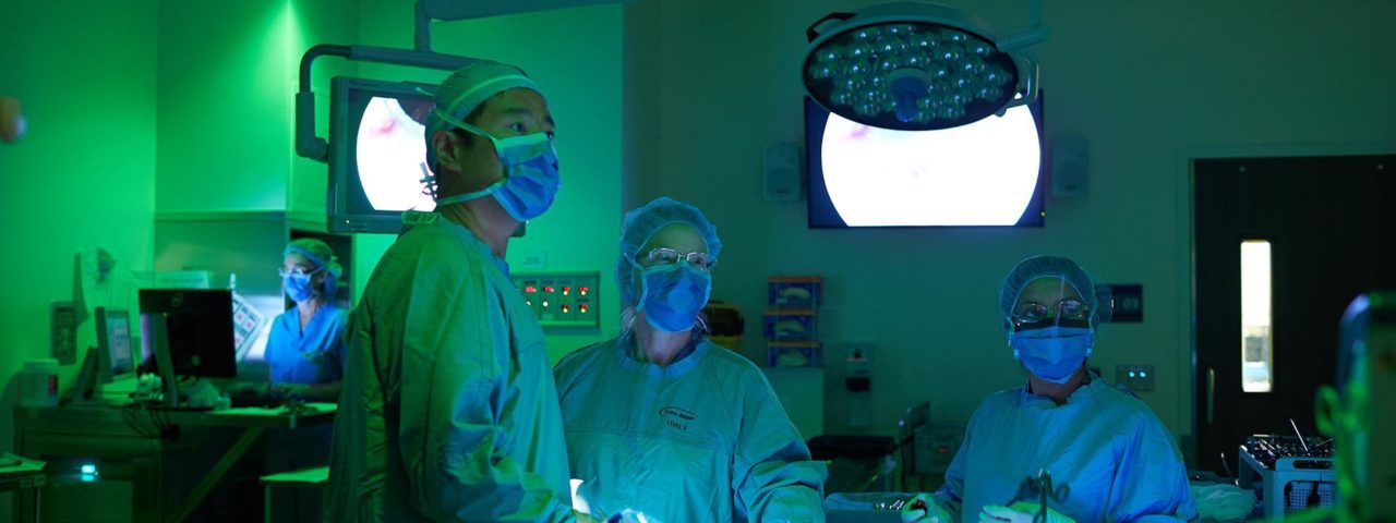 3 surgeons in a darkened operating room look in the same direction