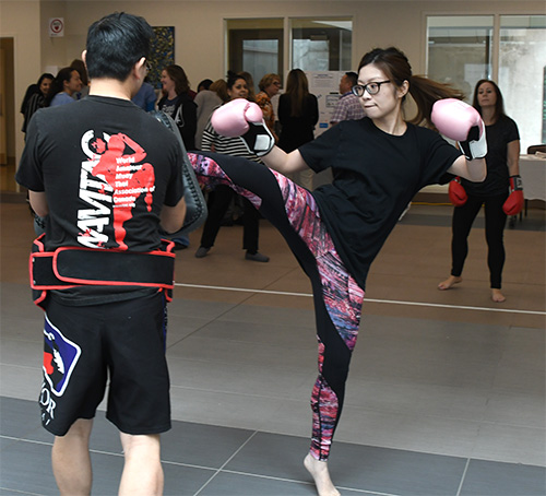 a woman in boxing gloves kicks up at an instructor holding a foam pad