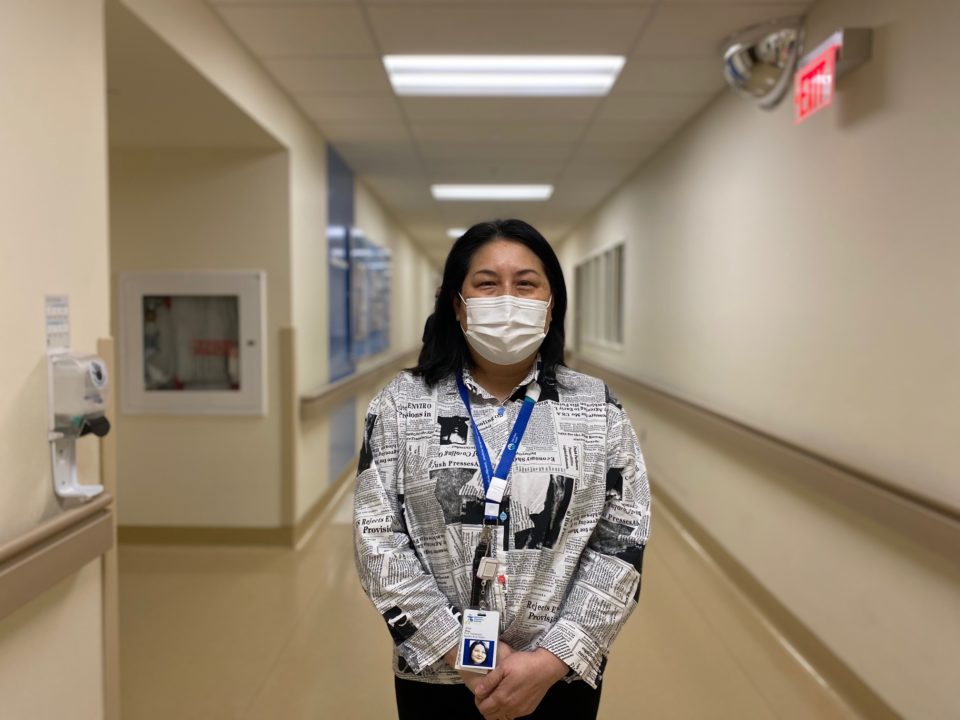 a woman in business clothes and medical mask stands in the hospital hallway and smiles for a photo