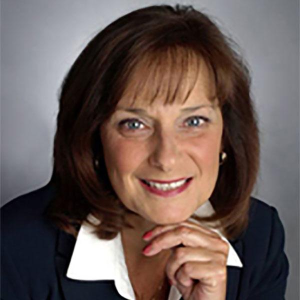 nancy sanders, seen from the shoulders up, wearing a black suit with shoulder length brown hair and smiling