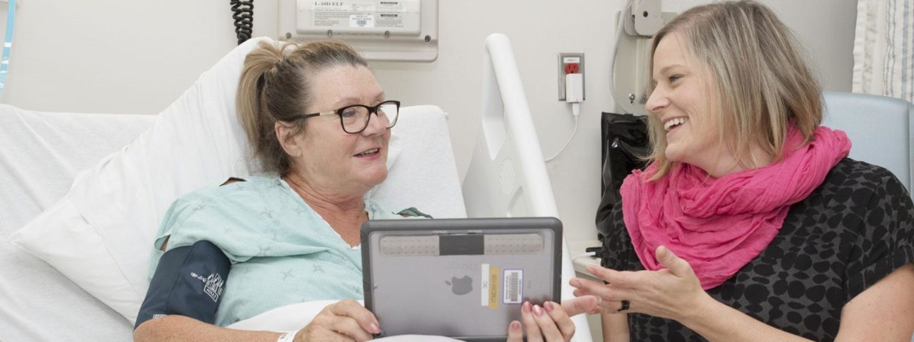 a female patient laying in a hospital bed holds an ipad and talks to a family member sitting next to her