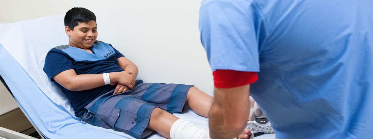 a smiling boy lays on a patient bed while an orthopedic doctor checks the cast on his right foot