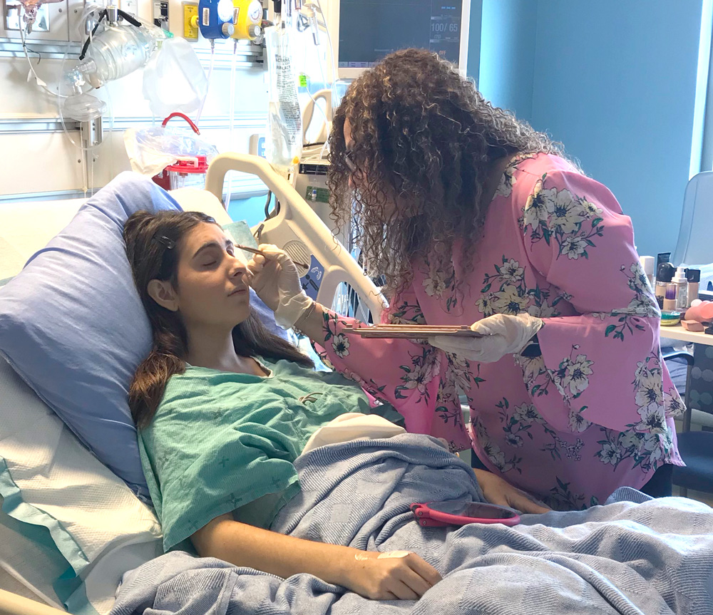 Elena lays in her hospital bed while another woman does her makeup