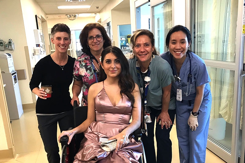 4 nurses stand behind Elena Papapetrou while she sits in her wheelchair wearing a pink prom dress