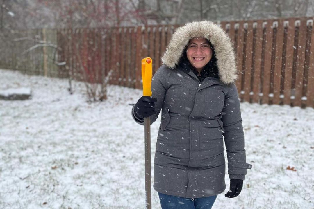 a woman in a grey coat standing in the snow and holding a shovel