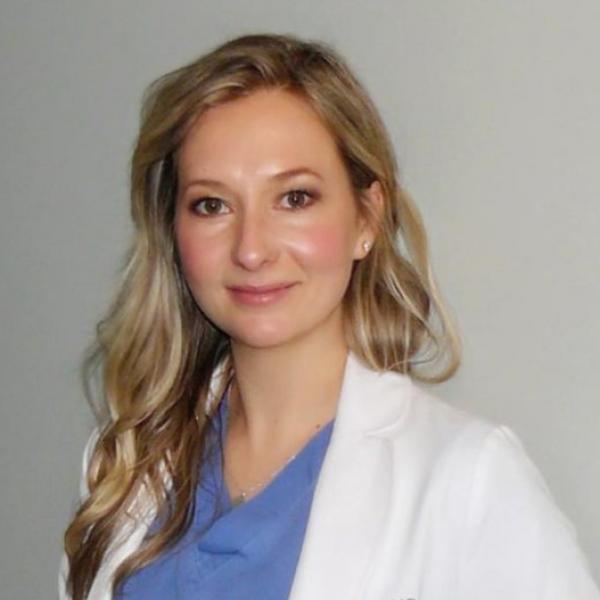 Dr. Victoria Hayward, seen from the chest up, wearing scrubs and a doctors coat, and long blond hair, smiling