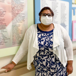 Dr. Deepa Grewal, seen from the waist up, wearing a patterned blue and white dress, white cardigan, glasses and medical mask