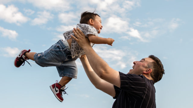 a father holds his toddler up in the air, both are smiling