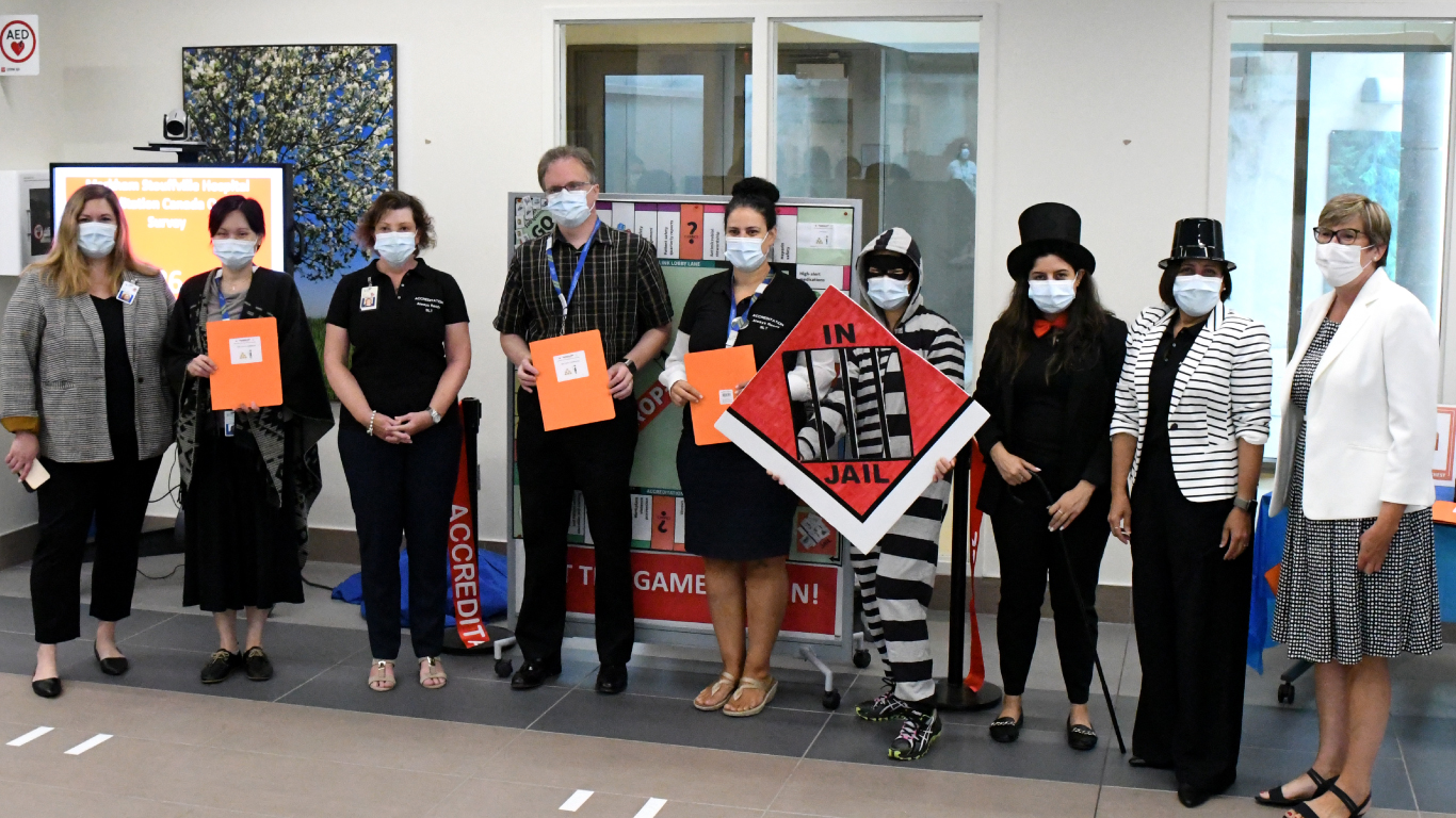 a group of men and women in business clothes and medical masks stand together with monopoly paraphernalia 