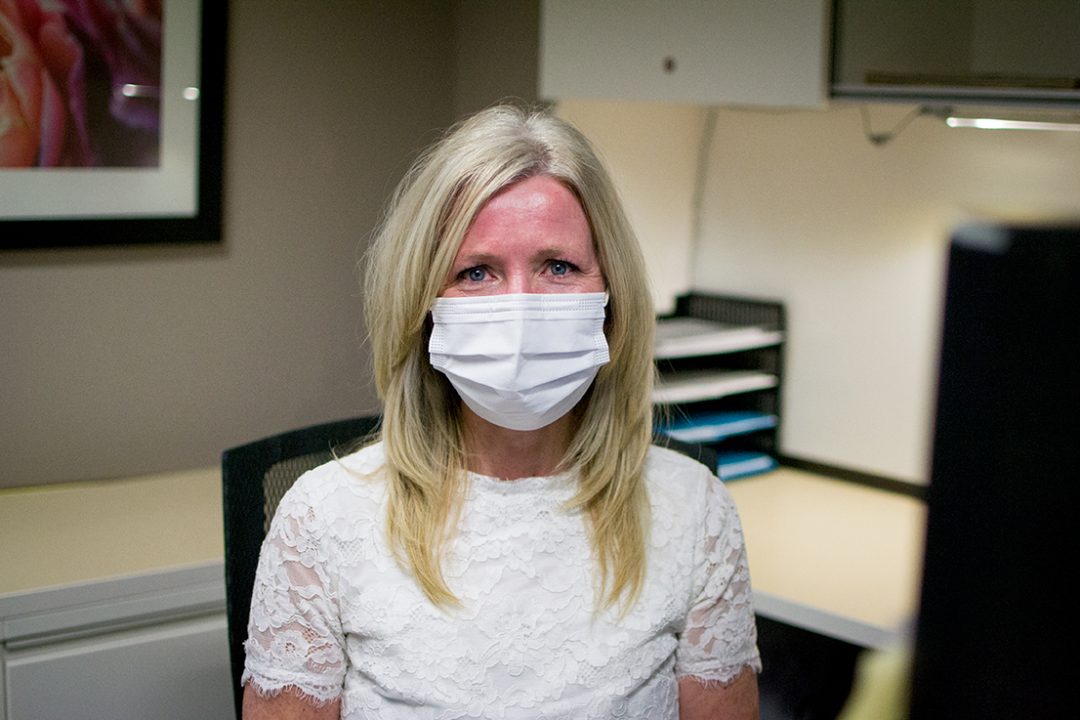 Megan Denby seen from the waist up sitting at her desk wearing a mask.
