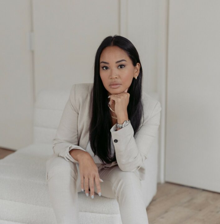 Esteena sits on a white chair wearing a white suit and her head resting on a closed fist.