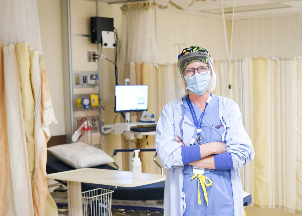 A nurse stands with their arms folded inside a hospital room wearing PPE.