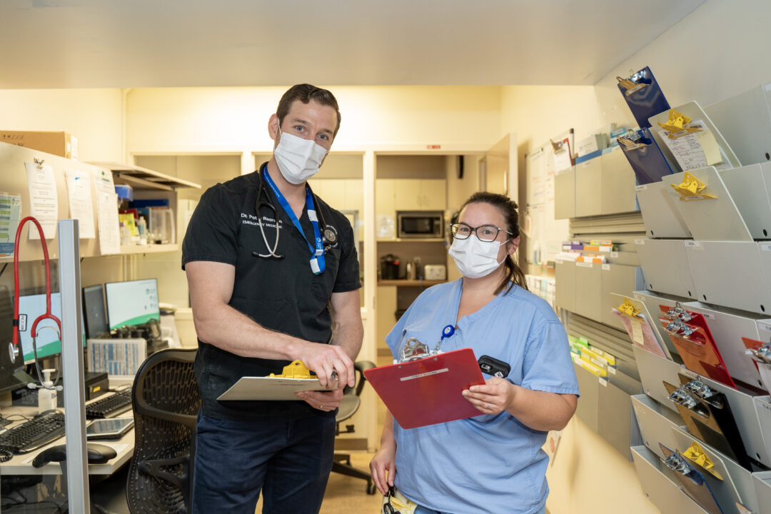 Two health care workers standing side-by-side inside a hospital wearing masks and holding clipboards.