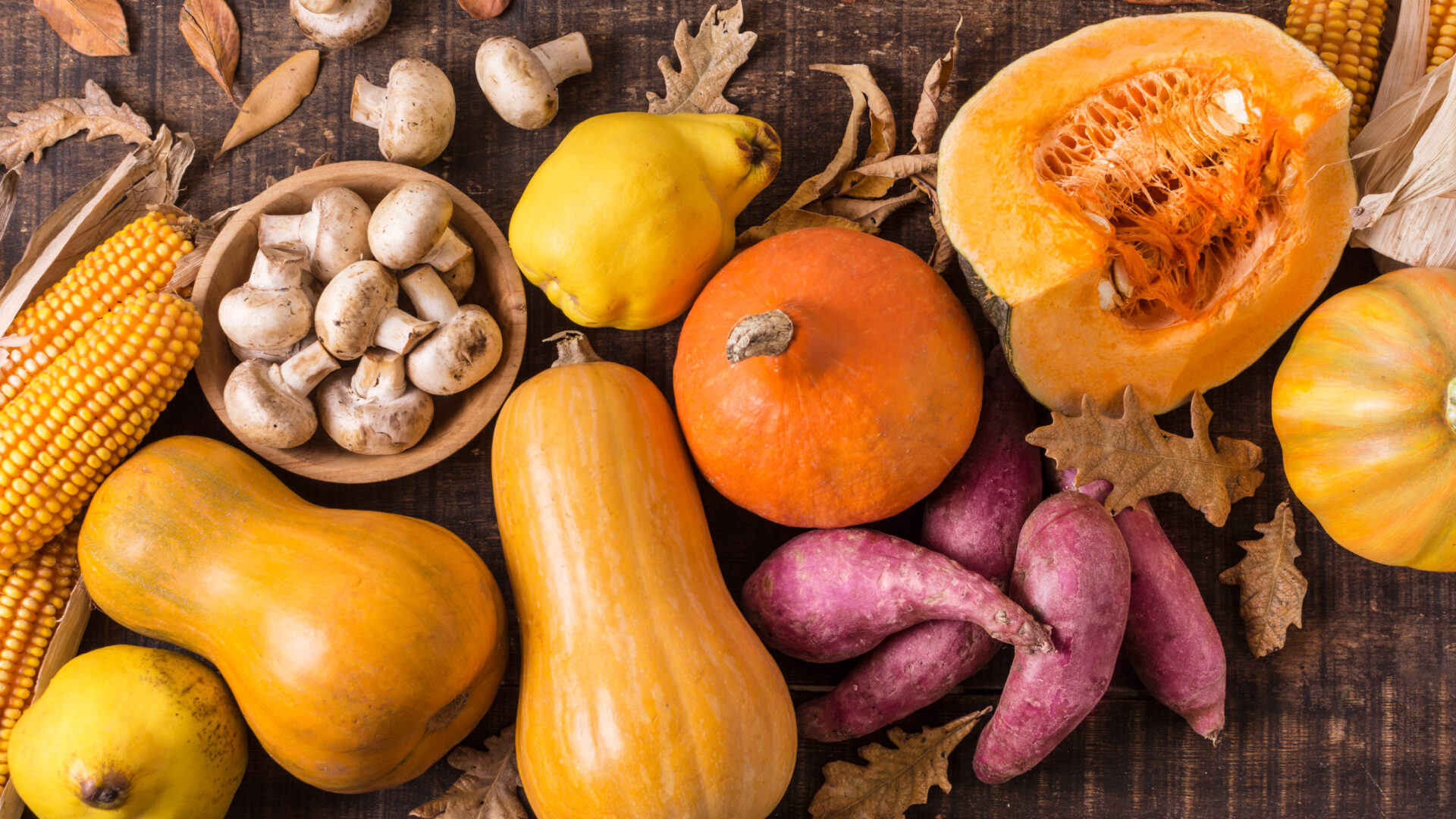 image of vegetables from the fall season such as pumpkins, corns, sweet potatoes and squash, nicely layout on top of wooden table with fallen leaves and mushrooms