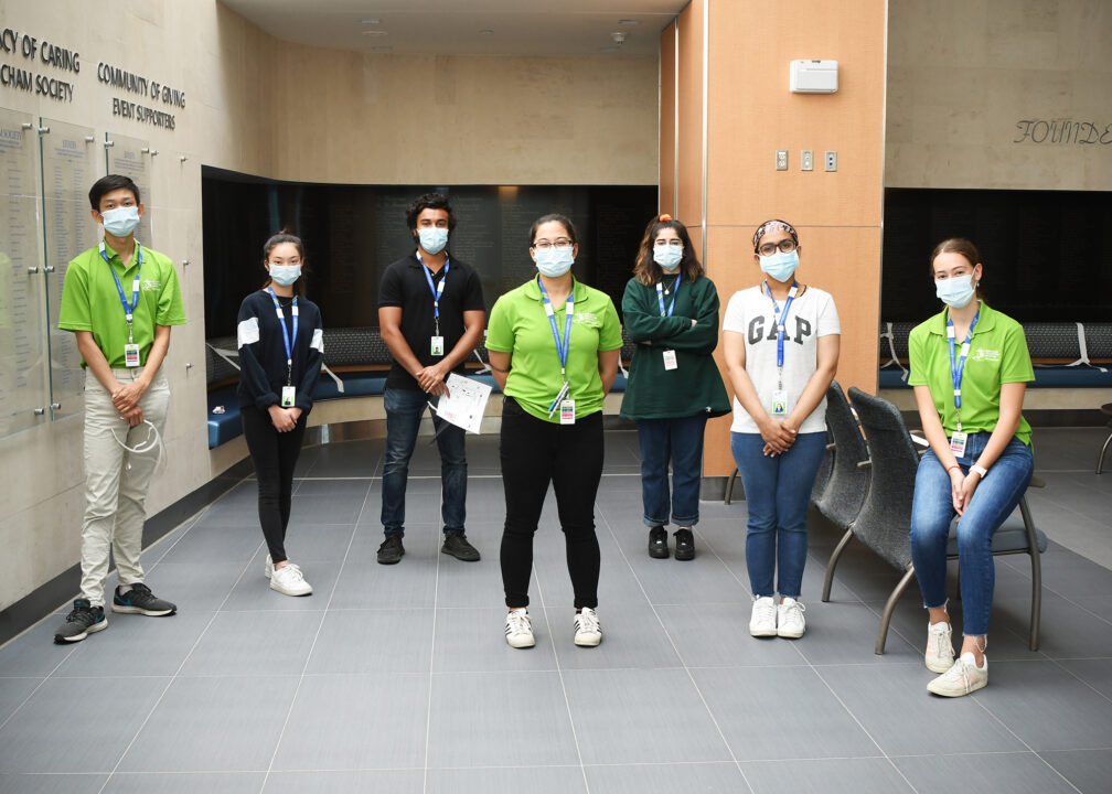 A group of volunteers wearing green shirts and masks stand spaced out inside a lobby at Markham Stouffville Hospital.