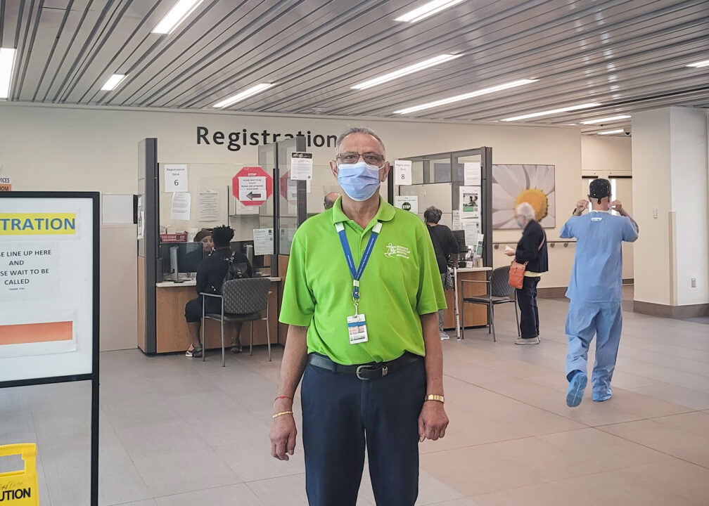 A hospital volunteer wearing a green shirt and a mask stands in front of the registration desk at Markham Stouffville Hospital.