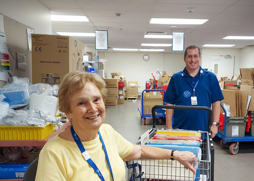 Two volunteers in a large storage room. One female sitting down wearing a yellow shirt. One male standing behind wearing a blue shirt.