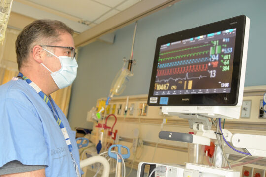 A hospital staff member stands next to and looks at the screen of a new Phillips monitor. He is standing next to the bed and wearing a mask.