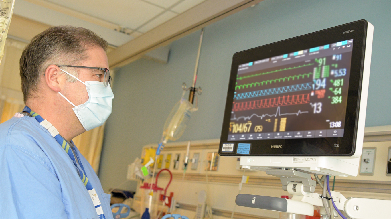 A hospital staff member stands next to and looks at the screen of a new Phillips monitor. He is standing next to the bed and wearing a mask.
