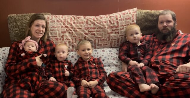 A mother and father snuggle on the couch with their four young children. All are wearing matching pajamas.