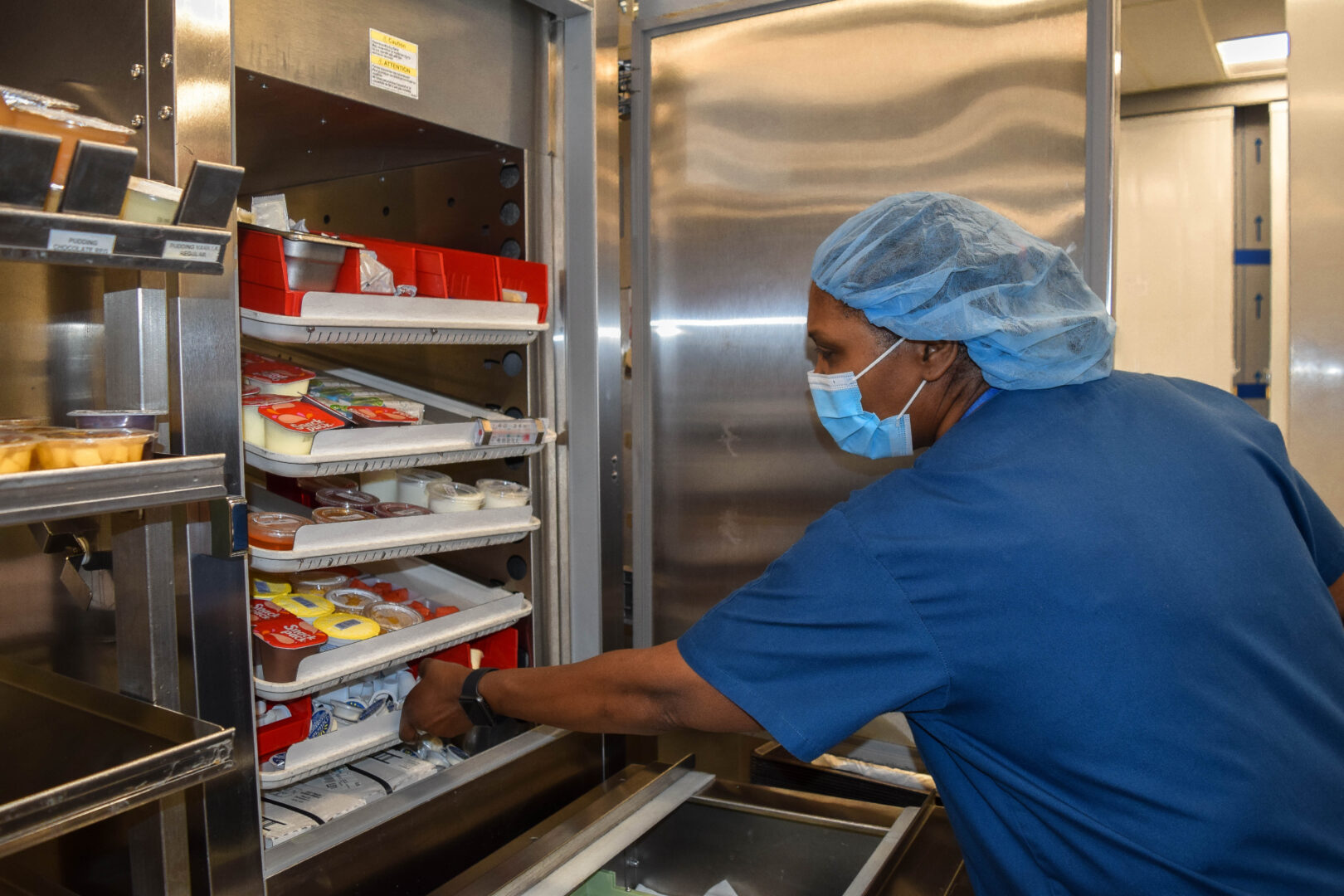 Photo of Karen Julien, a food service assistant at oak valley health, she is shown taking food compartments out of the fridge, she is wearing a face and hair mask in a blue gown