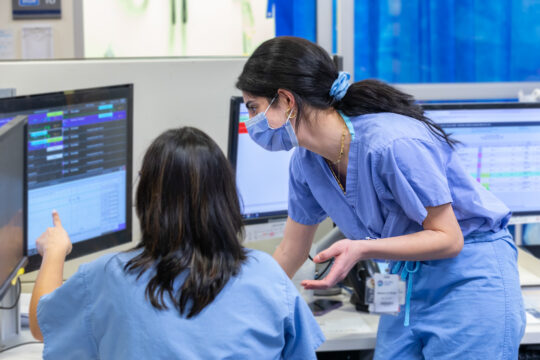 Two nurses stand together looking at a computer screen inside a hospital unit.